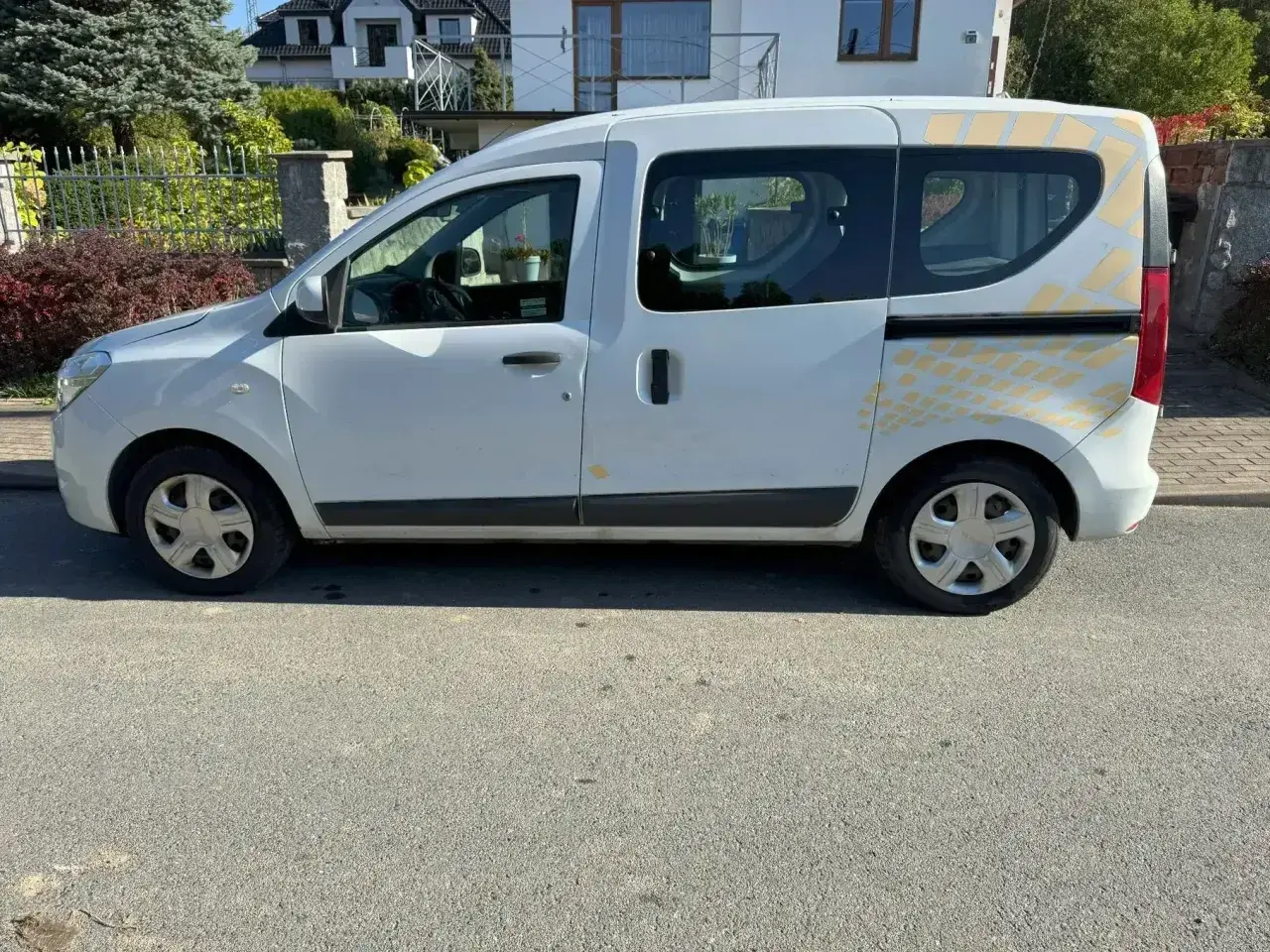 Biały Renault Dokker z żółtymi naklejkami na tylnej części, zaparkowany na ulicy.