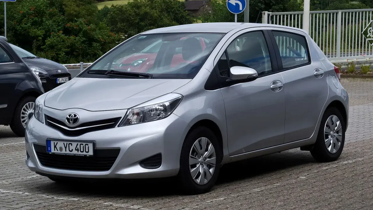 Srebrny Toyota Yaris P13 zaparkowany na kostce brukowej, z widocznym czerwonym samochodem w tle.