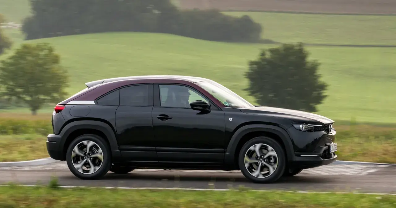 Czarna Mazda MX-30 z bordowym dachem sunie po drodze wśród zielonych wzgórz.