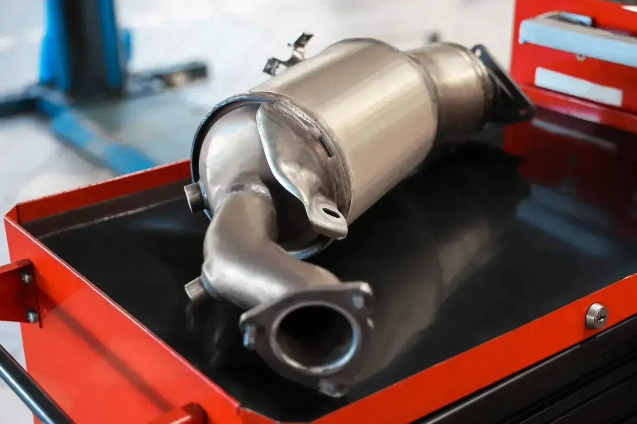 Metalowy element układu wydechowego samochodu, prawdopodobnie filtr DPF, leżący na wózku warsztatowym.