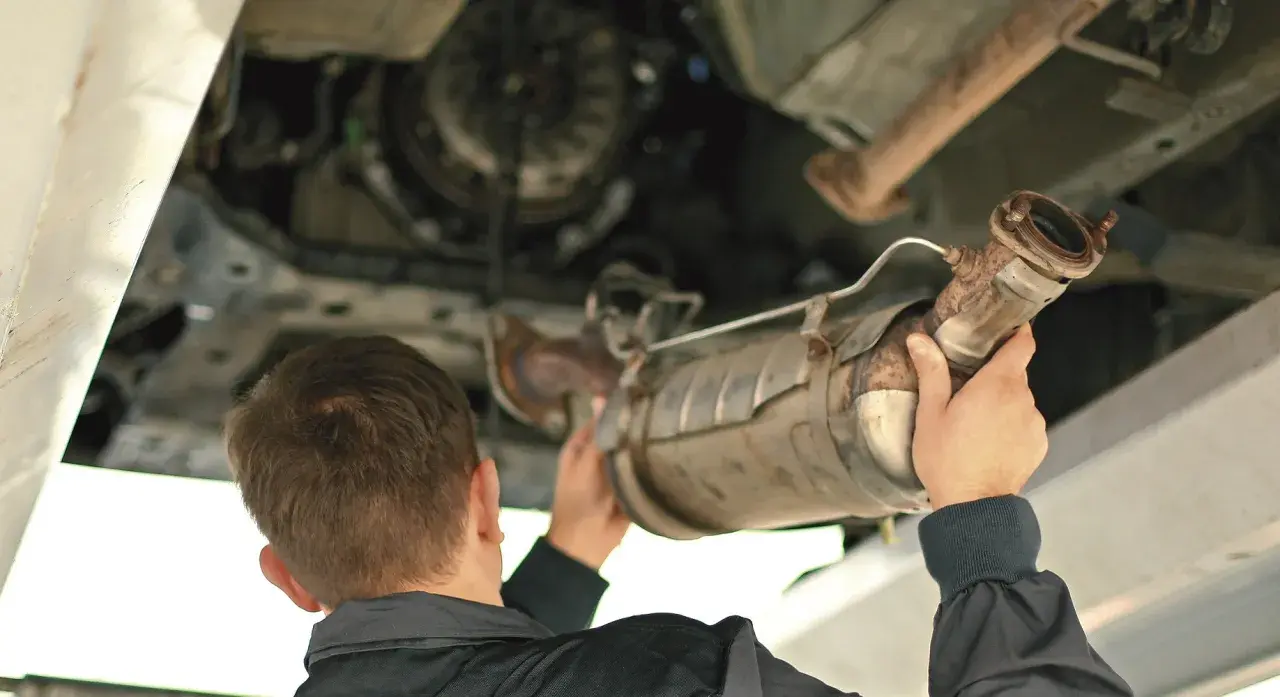 Mechanik trzymający pod samochodem zardzewiały element układu wydechowego, prawdopodobnie DPF.