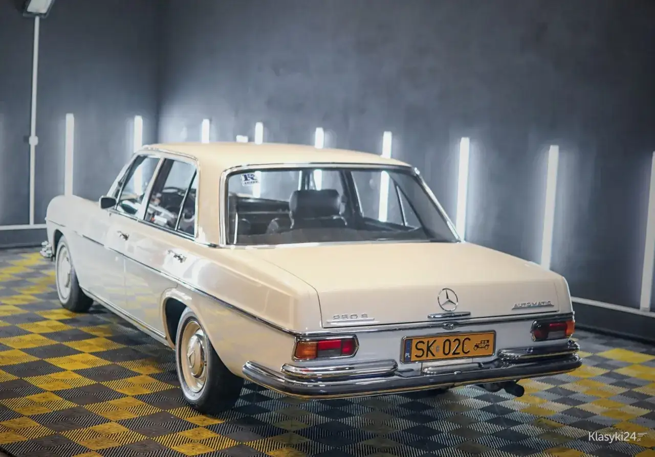 Biały Mercedes 280S z lat 70. w104, lśniący na tle nowoczesnych lamp. Klasyka motoryzacji.
