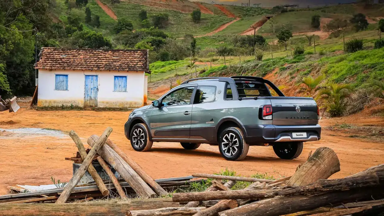 Szary Volkswagen Saveiro Extreme na wiejskiej drodze, obok małego domu z niebieskimi okiennicami.
