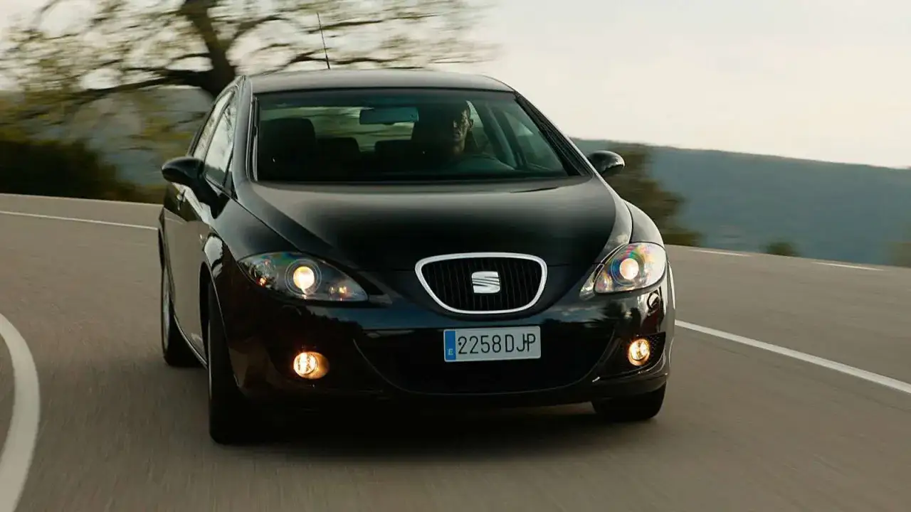 Czarny Seat Leon 1P1 na zakręcie drogi, światła włączone, kierowca za kierownicą.