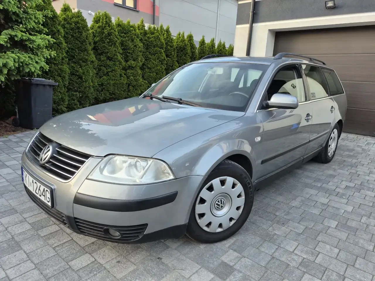 Szary Volkswagen Passat B5.5 kombi stoi na podjeździe z kostki brukowej, otoczony tujami.