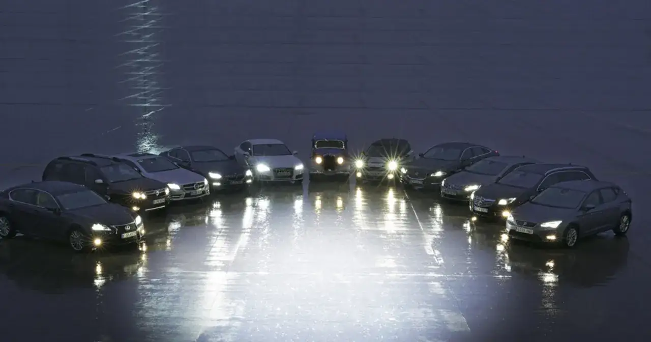 Rząd samochodów z włączonymi światłami mijania i dziennymi, tworząc refleksy na mokrej nawierzchni.