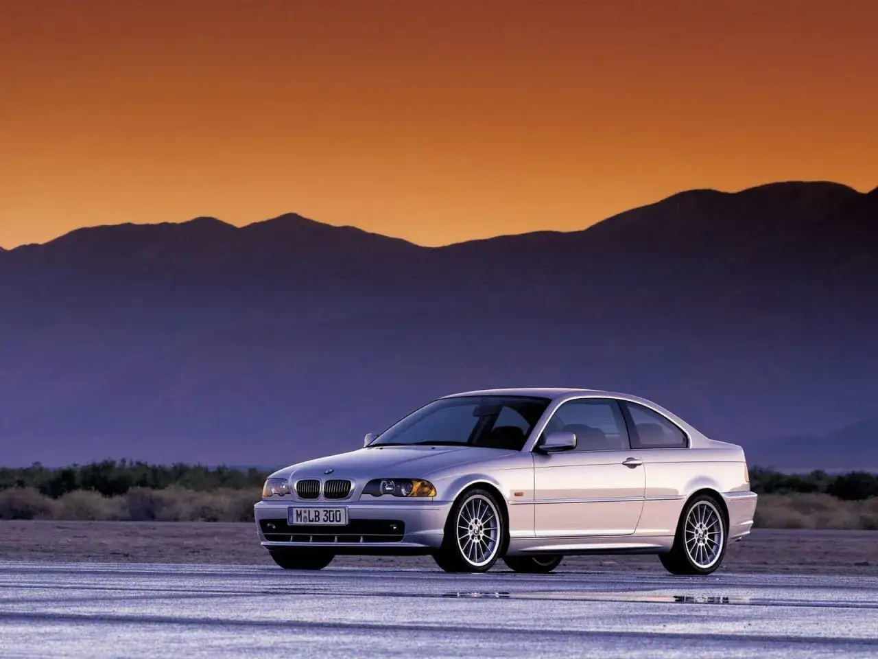 Srebrne BMW E46 coupe na pustynnej drodze o zachodzie słońca. Ten model, z charakterystycznym rokiem produkcji, prezentuje się elegancko na tle gór.