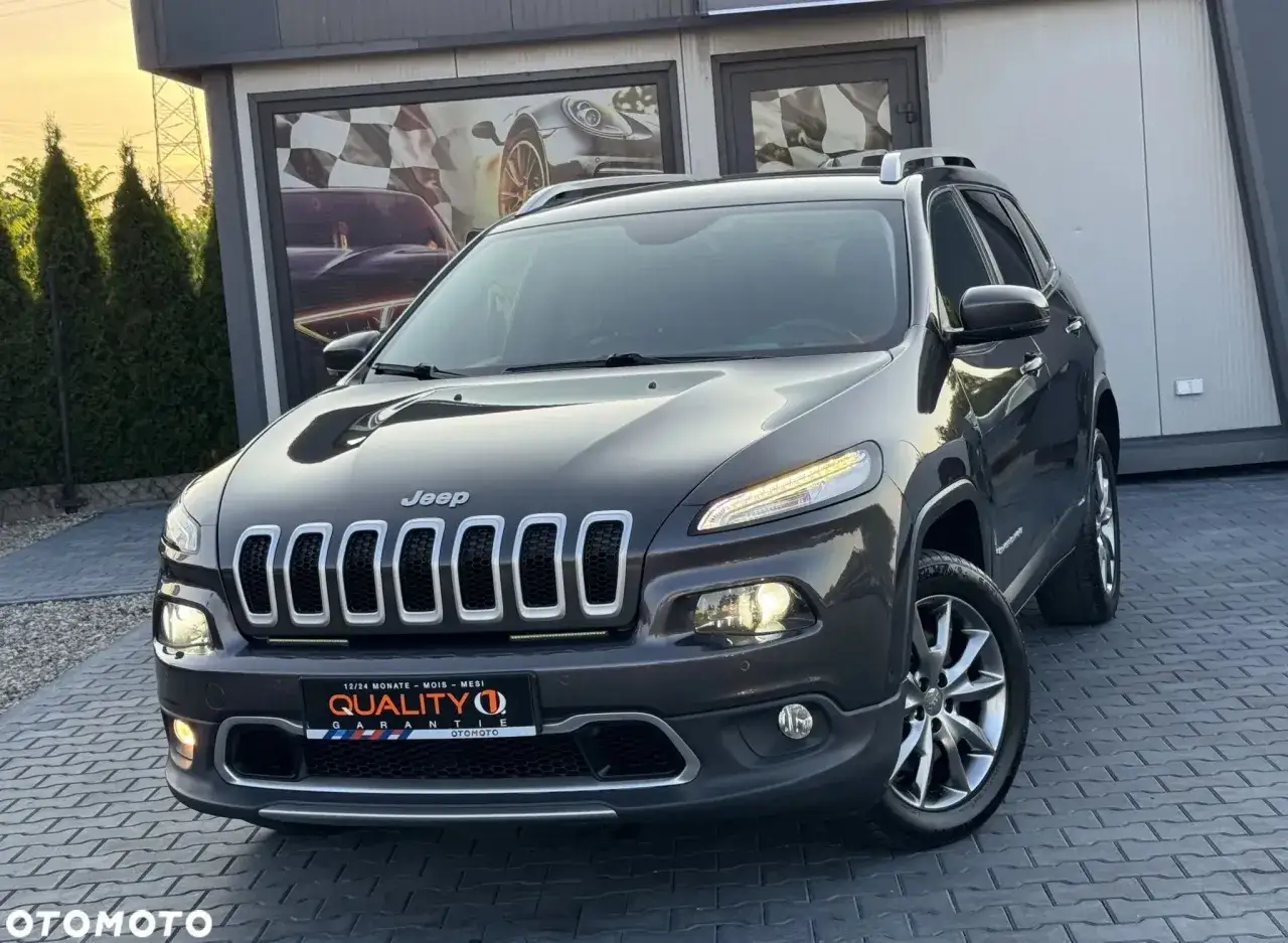 Ciemnoszary Jeep Cherokee KL z chromowanymi elementami, zaparkowany przed budynkiem z motywami wyścigowymi.