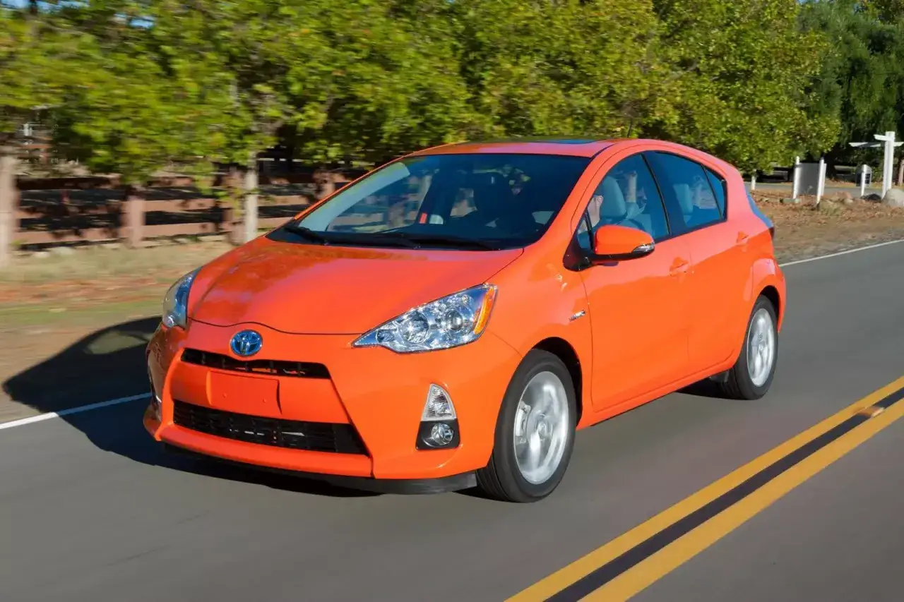 Pomarańczowy Prius C sunie po drodze wśród zieleni.