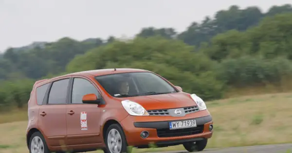 Pomarańczowy Nissan Note E12 z kobietą za kierownicą jedzie po drodze wśród zieleni.