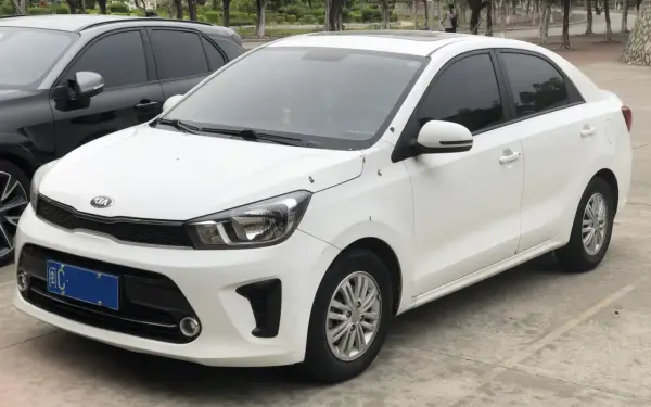Biały sedan Kia Pegas z przyciemnianymi szybami zaparkowany obok czarnego SUV-a.