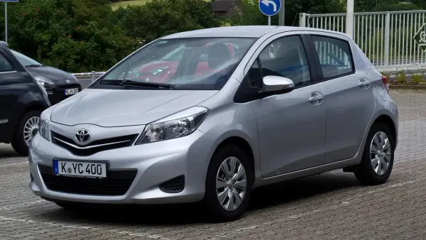 Srebrny Toyota Yaris P13 zaparkowany na kostce brukowej, z widocznym czerwonym samochodem w tle.