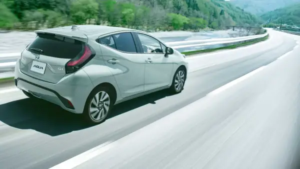 Jasnoszary Toyota Aqua jedzie po krętej drodze wśród zielonych wzgórz.